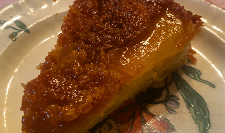 Part de gâteau aux pommes caramélisées servie sur une assiette fleurie ancienne, posée sur une nappe rose. La texture dorée du gâteau et sa surface brillante de caramel révèlent une cuisson maison soignée, typique d’un gâteau renversé aux pommes. L’ambiance évoque un moment de dégustation chaleureux et authentique, parfait pour un dessert convivial.