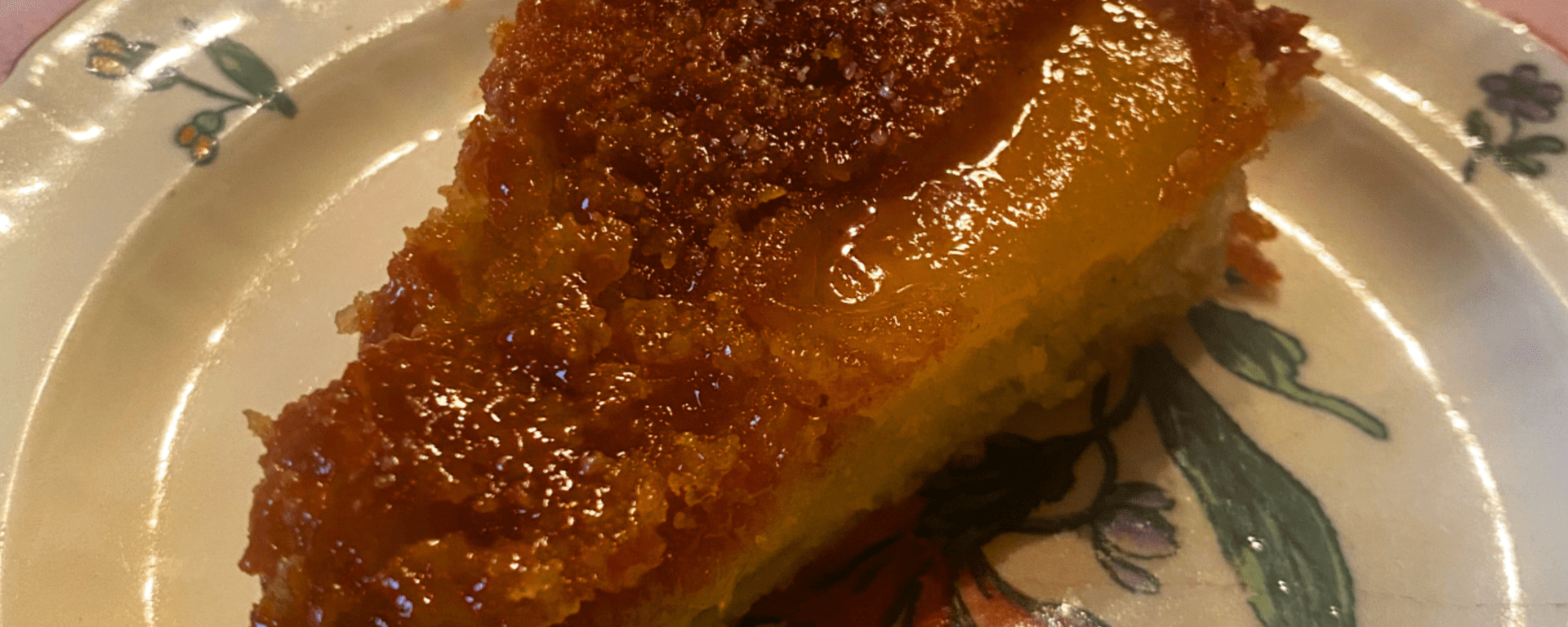 Part de gâteau aux pommes caramélisées servie sur une assiette fleurie ancienne, posée sur une nappe rose. La texture dorée du gâteau et sa surface brillante de caramel révèlent une cuisson maison soignée, typique d’un gâteau renversé aux pommes. L’ambiance évoque un moment de dégustation chaleureux et authentique, parfait pour un dessert convivial.